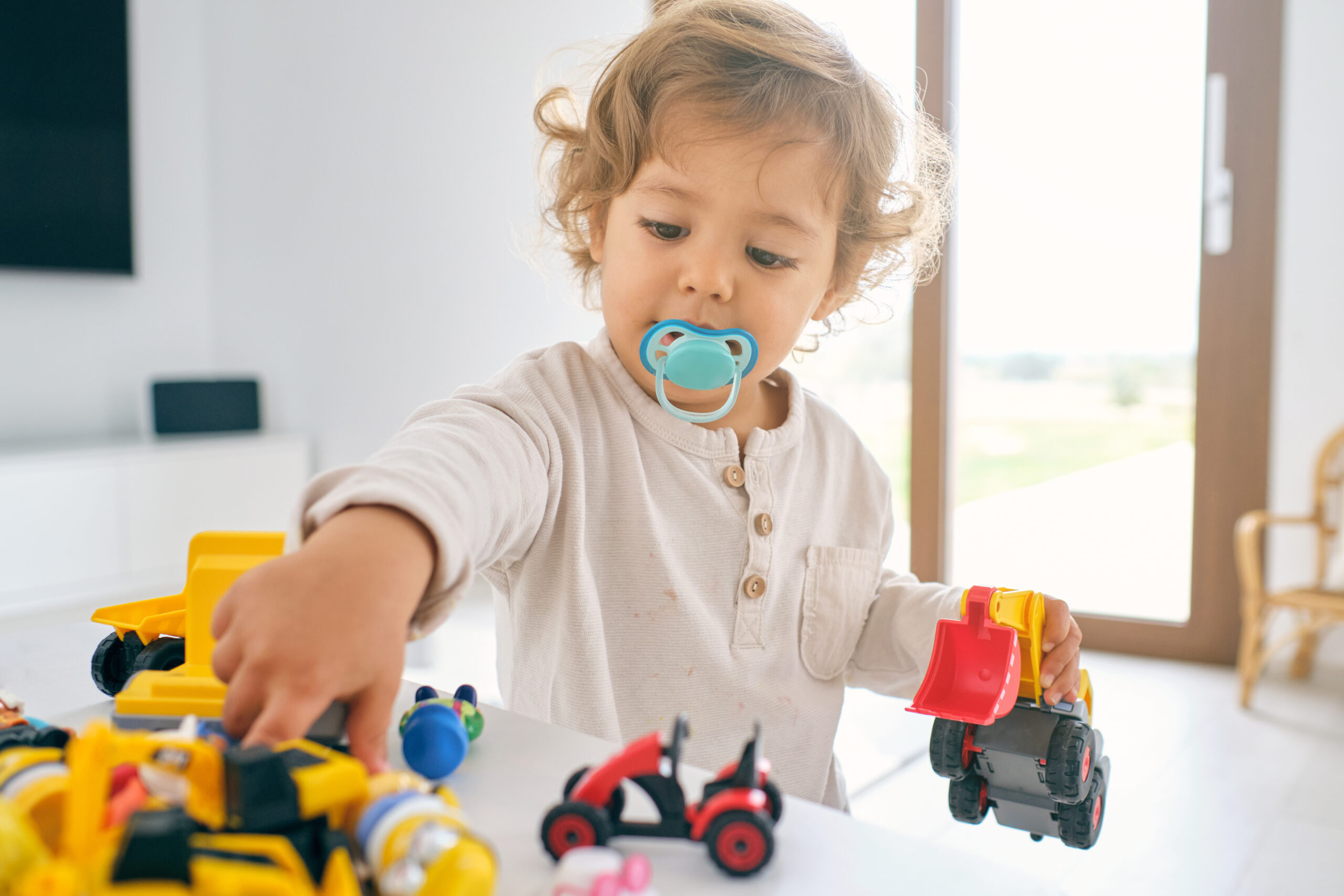 Adorable little child with pacifier playing with colorful plastic toys at home in sunny day on blurred background